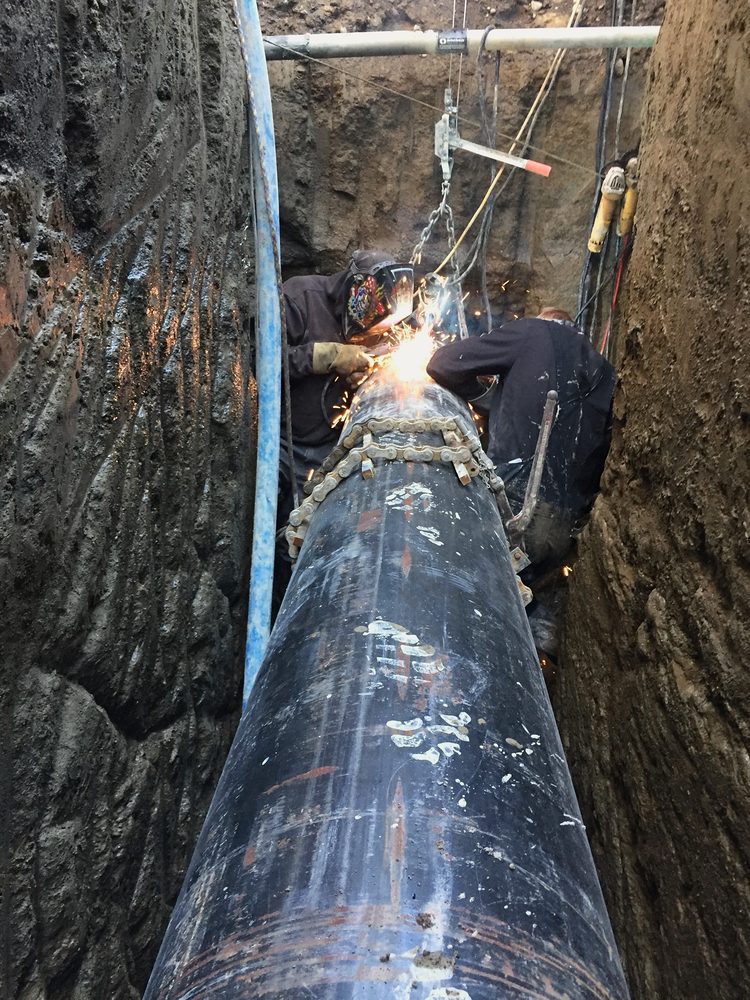 Welders working down in a trench on a large-diameter steel pipe, arc glow, crew in hoods