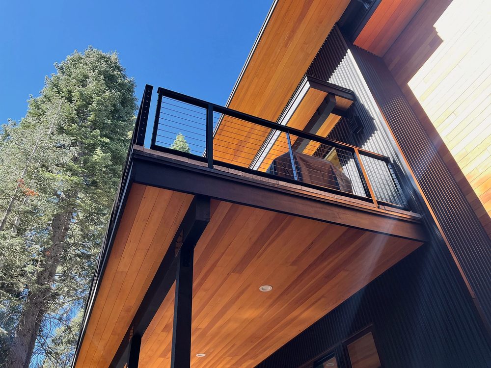 Looking up at a cantilevered balcony with black steel post and cable railing, cedar soffit, black siding, tall pine and blue sky