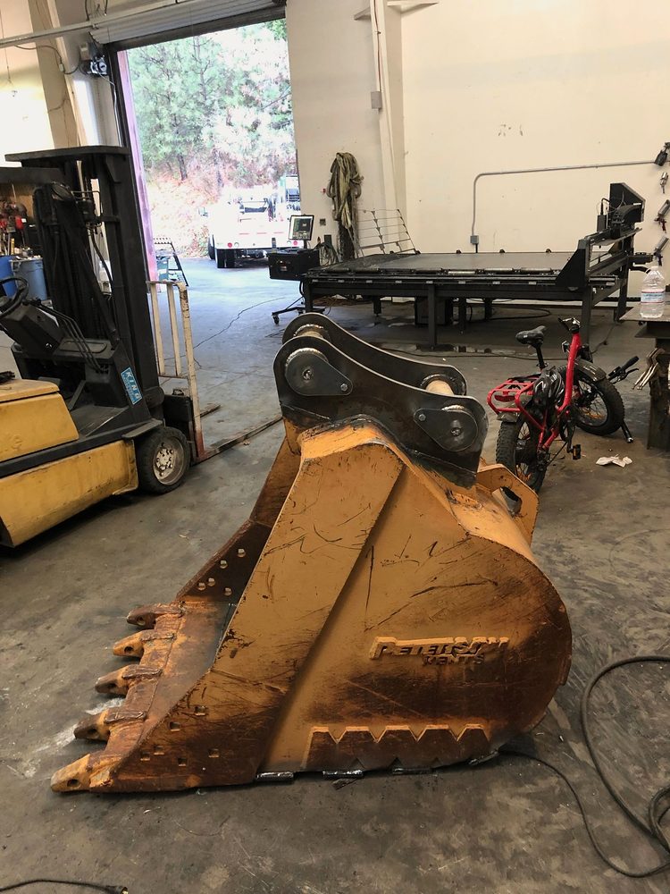 Excavator bucket repair on shop floor, forklift, flatbed and roll-up door behind