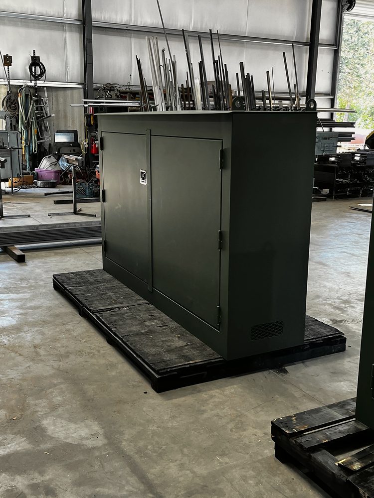 Dark green powder-coated steel equipment enclosure on a pallet inside the shop, stock racks and machines behind